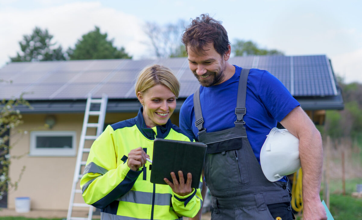 A man and woman solar installers engineers with tablet while installing solar panel system on house.